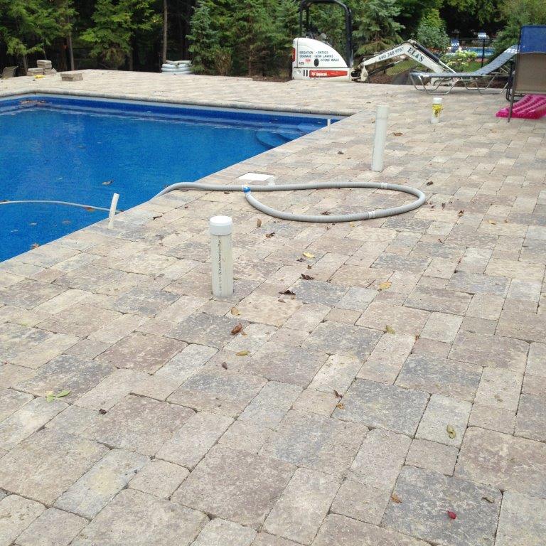 Pool deck with stone pavers, blue swimming pool, and a coiled hose on the ground.