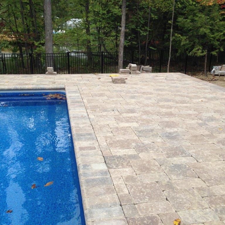 Stone patio beside a blue swimming pool, bordered by trees and a black fence.