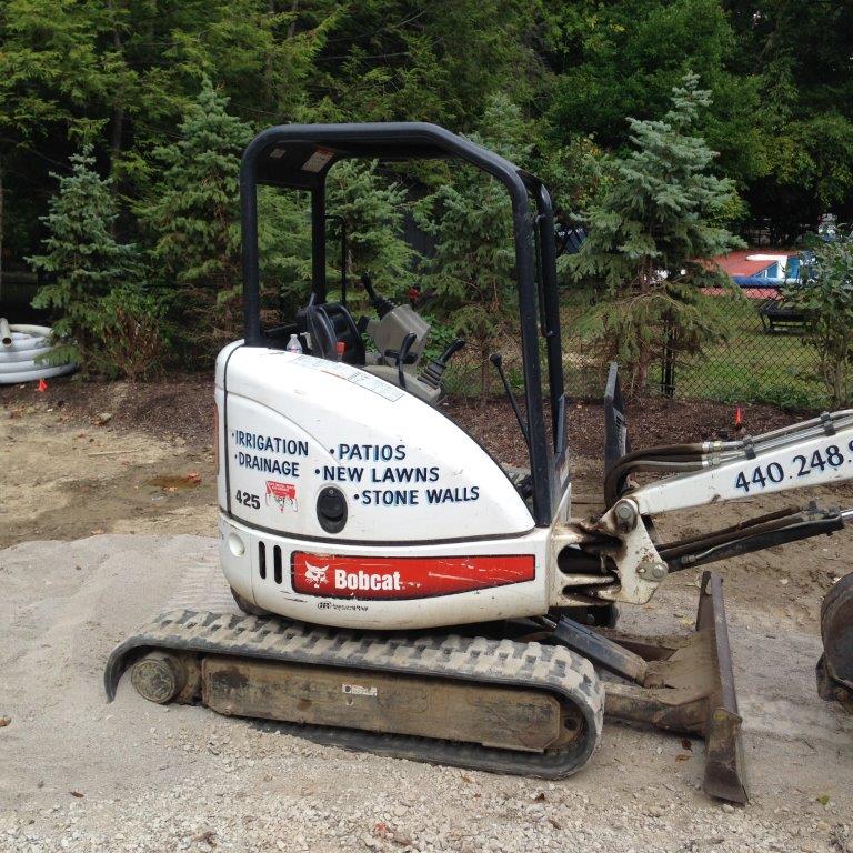 Small Bobcat excavator on a dirt lot beside trees, with a bucket and company decals visible.