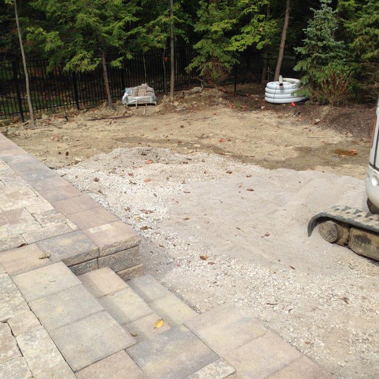Backyard with stone patio, bare dirt area, and stacked metal fire pits near a wooded fence