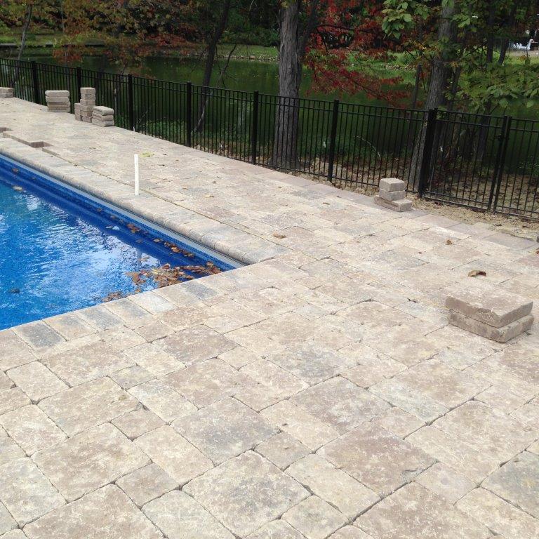 Stone pool deck beside blue swimming pool, with black fence and trees in the background.