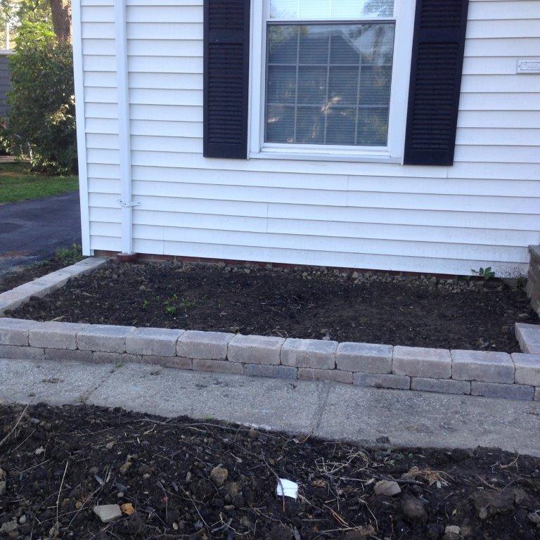 Front of a white house with a window, black shutters, and a raised garden bed bordered by stone blocks.
