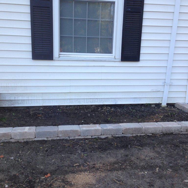 White house siding with a window, black shutters, and a stone border along a bare mulch bed