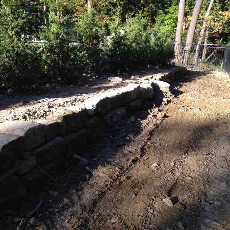 Stone retaining wall beside a dirt path in a sunlit wooded area