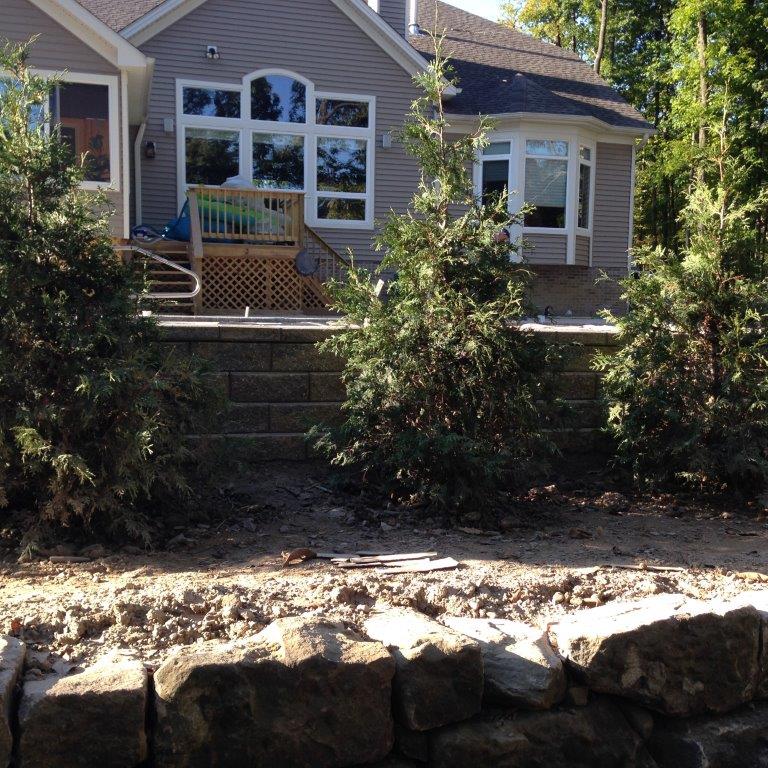 Brick house with large windows, front deck, and shrubs above a stone retaining wall.