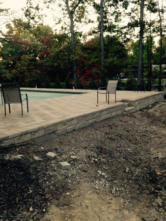 Patio with two chairs beside an empty pool, bordered by trees and a rough dirt foreground
