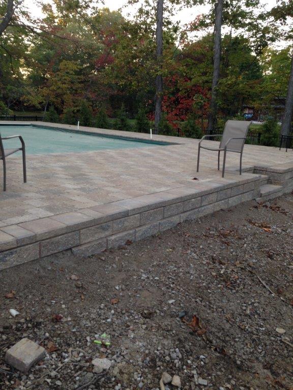 Patio beside an empty pool with chairs and a gravel foreground, backed by trees.