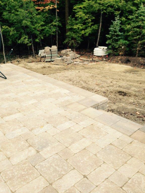 Stone patio beside a bare dirt yard, with trees and landscaping materials along the back edge.