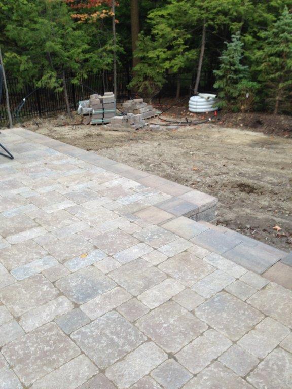 Stone patio beside a bare dirt yard with trees and landscaping materials in the background