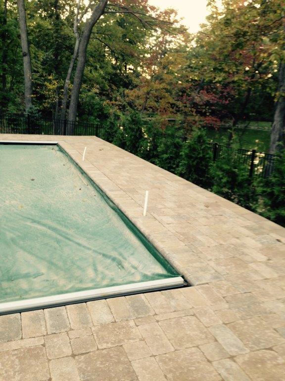Covered pool beside beige stone patio, with trees and greenery in the background