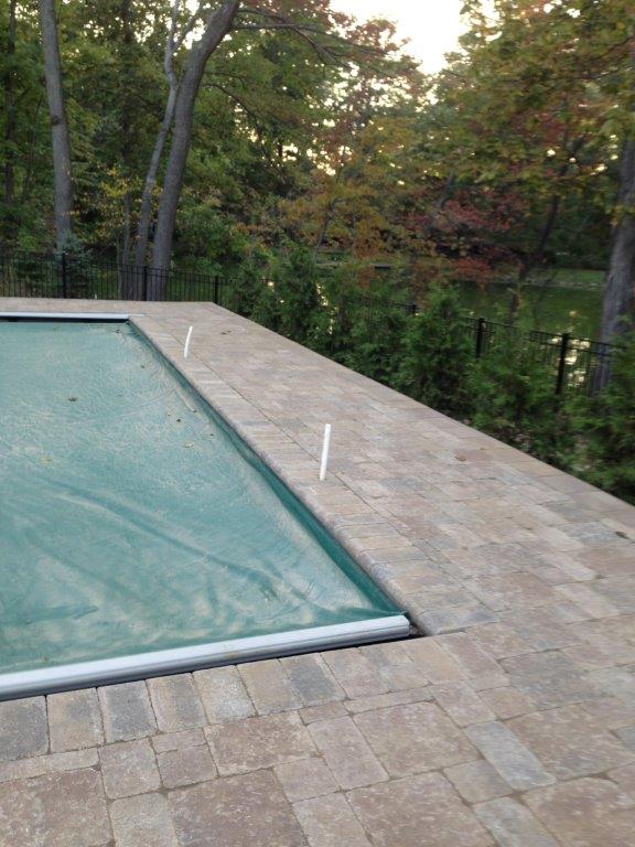 Pool with safety cover beside a stone patio and wooded backyard.
