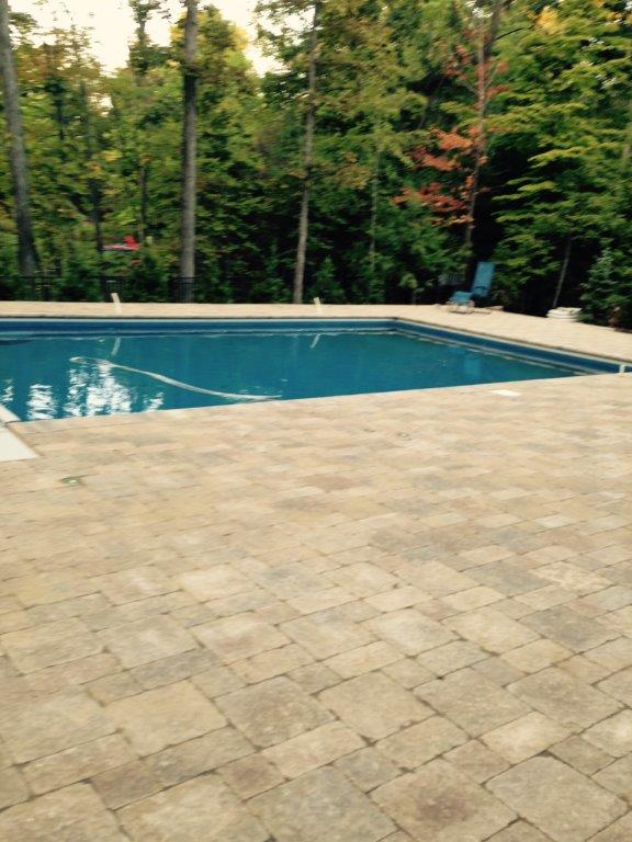Outdoor swimming pool beside a tan stone patio, bordered by trees and lounge chairs.