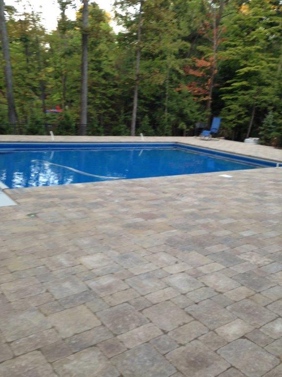 Backyard swimming pool beside a tan stone patio, surrounded by trees and lounge chairs
