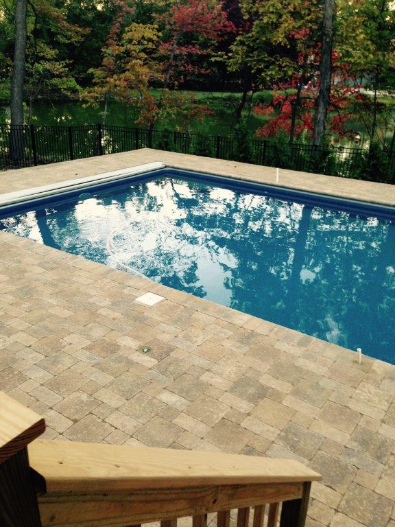 Backyard swimming pool with blue water beside a tan stone patio and autumn trees.