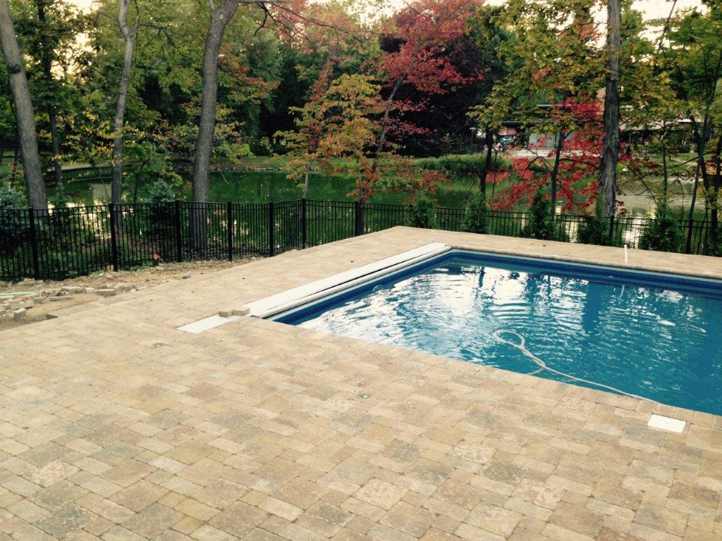 Backyard swimming pool beside a tan paver patio, with trees and a fence in the background