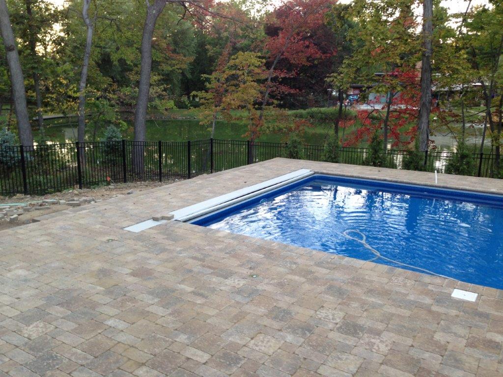 Outdoor pool with blue water beside a stone patio and trees beyond a black fence