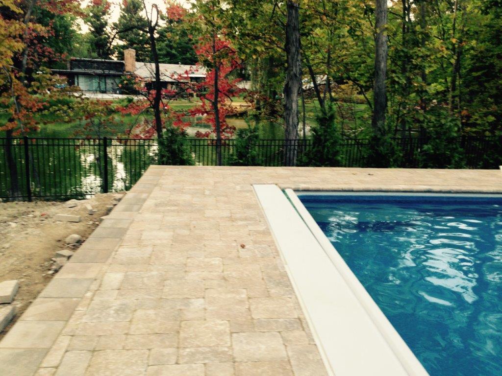 Stone patio beside a blue swimming pool with trees and a fenced garden in the background