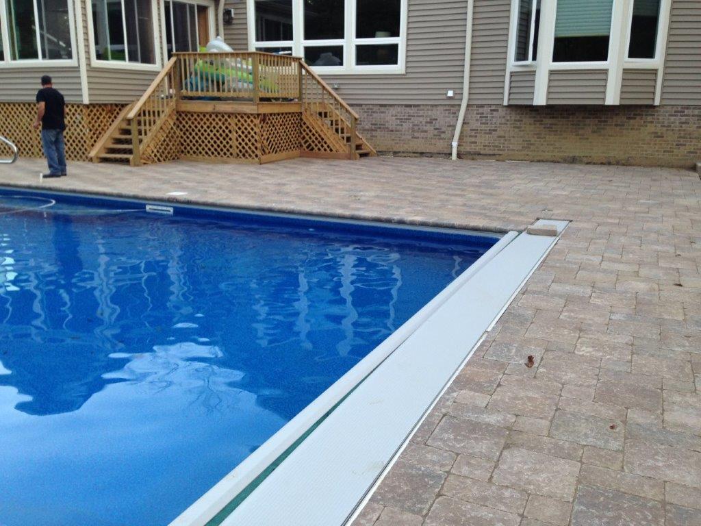 Backyard swimming pool beside a tan paver deck and house, with a child standing near the steps