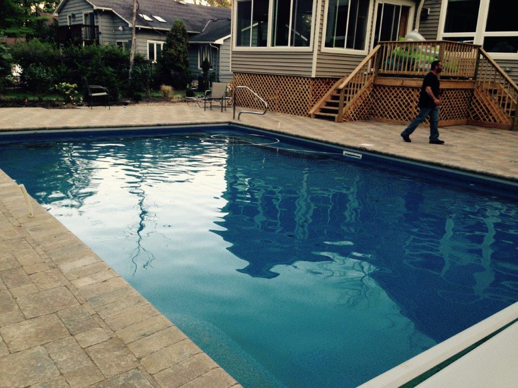 Backyard swimming pool with clear blue water beside a deck and house, with a person walking nearby