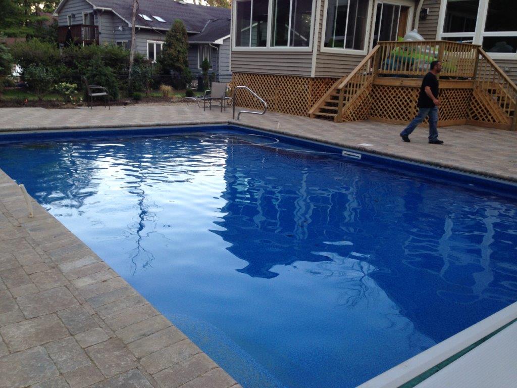 Backyard swimming pool with blue water, brick deck, and a person walking near a wooden deck staircase
