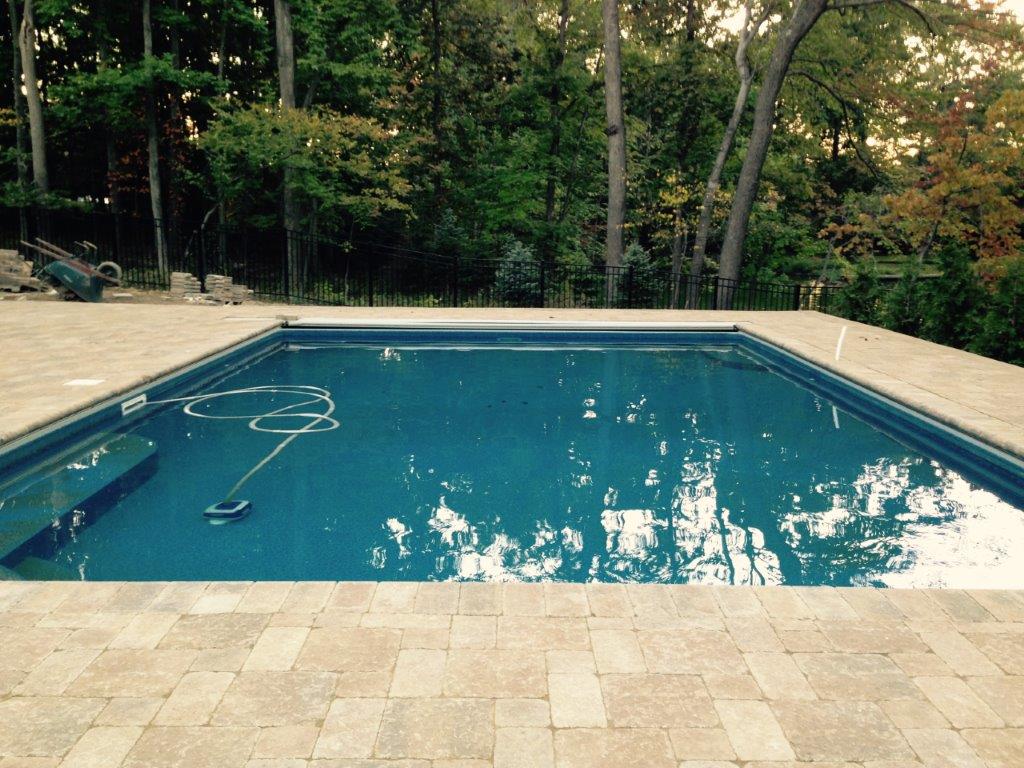Rectangular backyard swimming pool with blue water, tan stone deck, and trees in the background