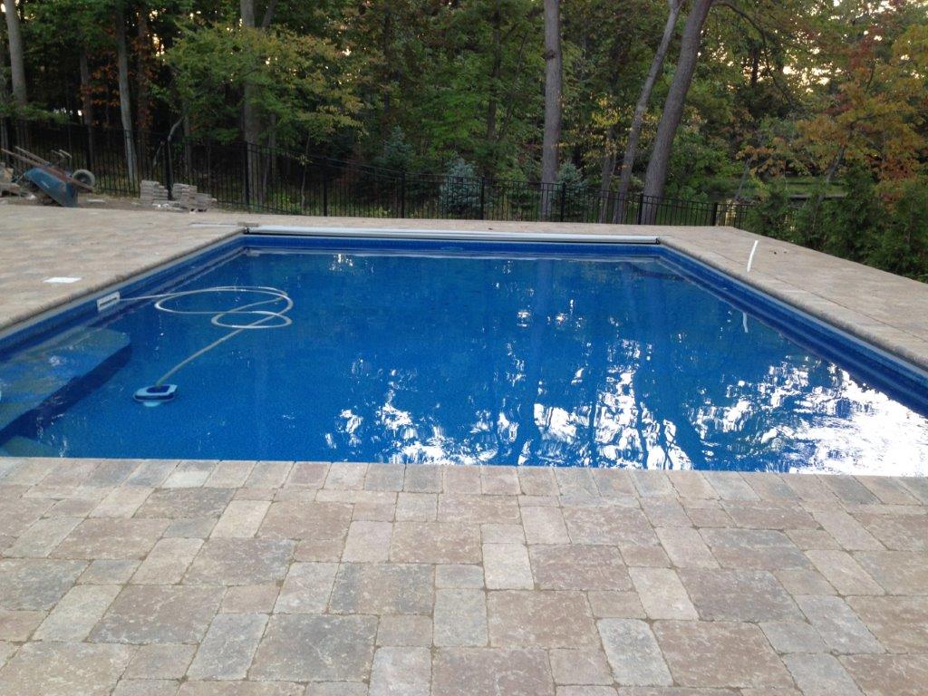 Rectangular blue swimming pool surrounded by stone patio and trees beyond