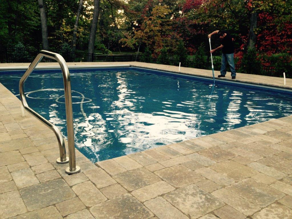 Outdoor swimming pool with blue water, stone deck, and metal handrail, bordered by trees and shrubs.