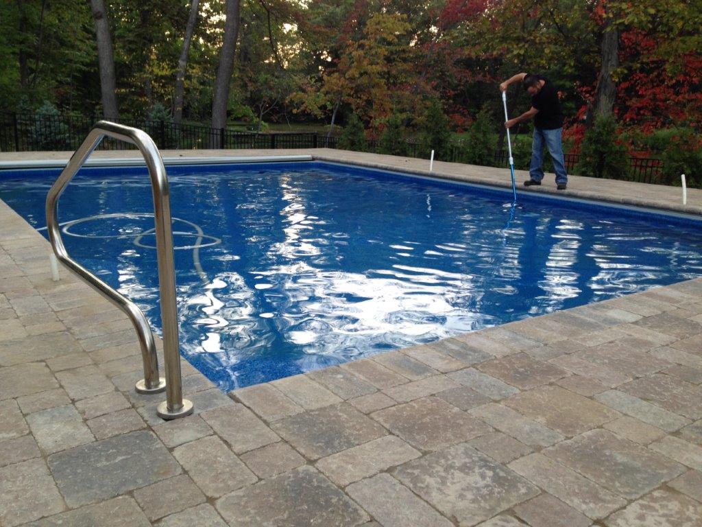 Empty blue swimming pool with a metal handrail and pavers, outdoors beside trees and a fence