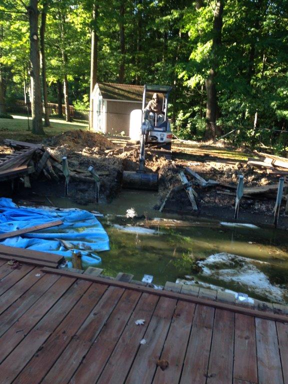 Dock beside a debris-filled pond with a small skid steer in a wooded area
