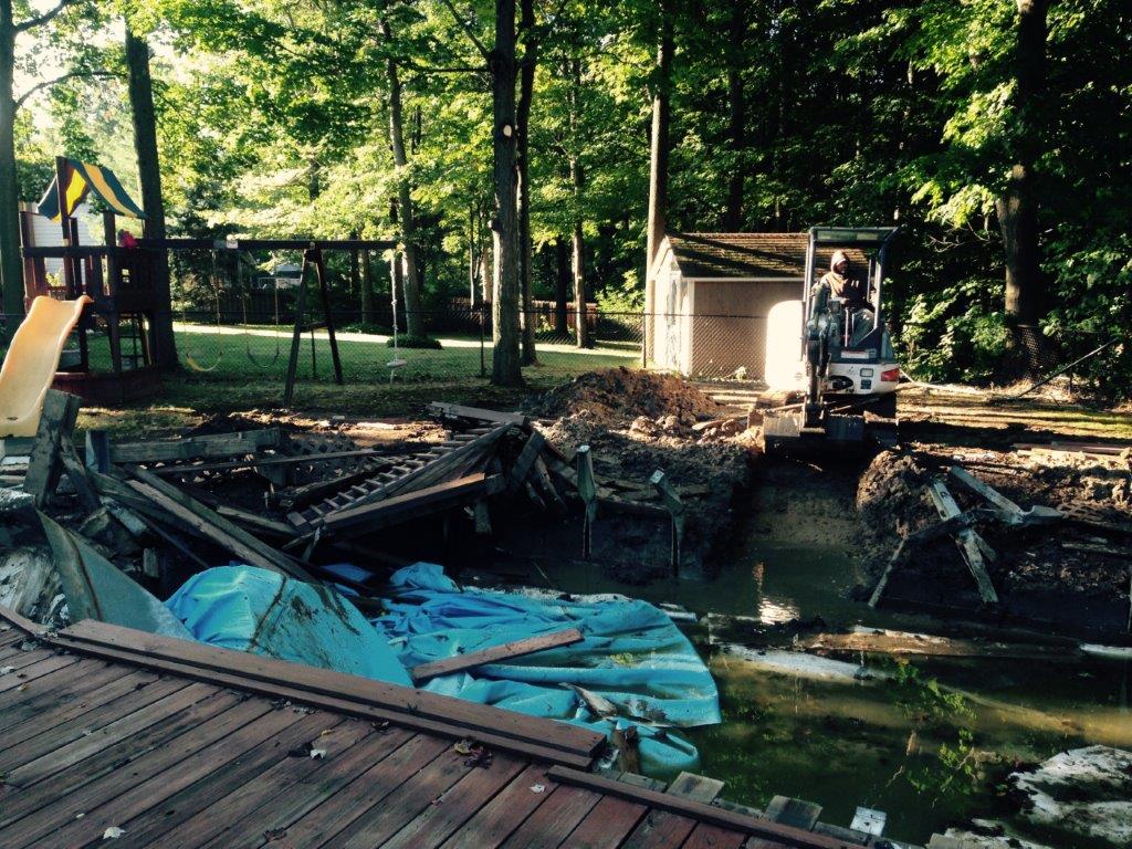 Damaged dock and debris by a wooded pond, with a small excavator clearing rubble
