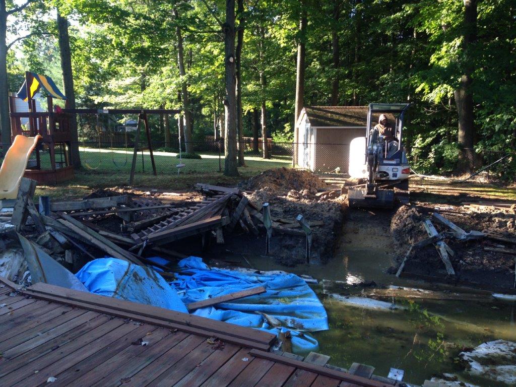 Fire-damaged backyard with a small excavator, charred debris, blue tarp, and trees nearby