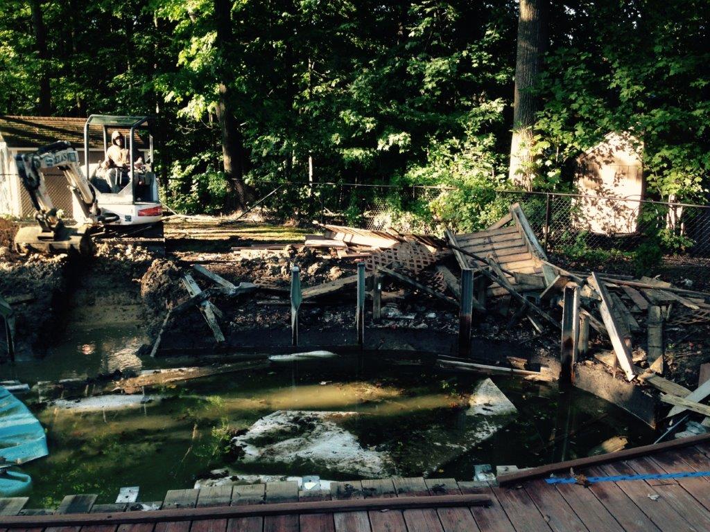 Collapsed wooden bridge over a shallow creek in a wooded area, with rubble and a small vehicle nearby