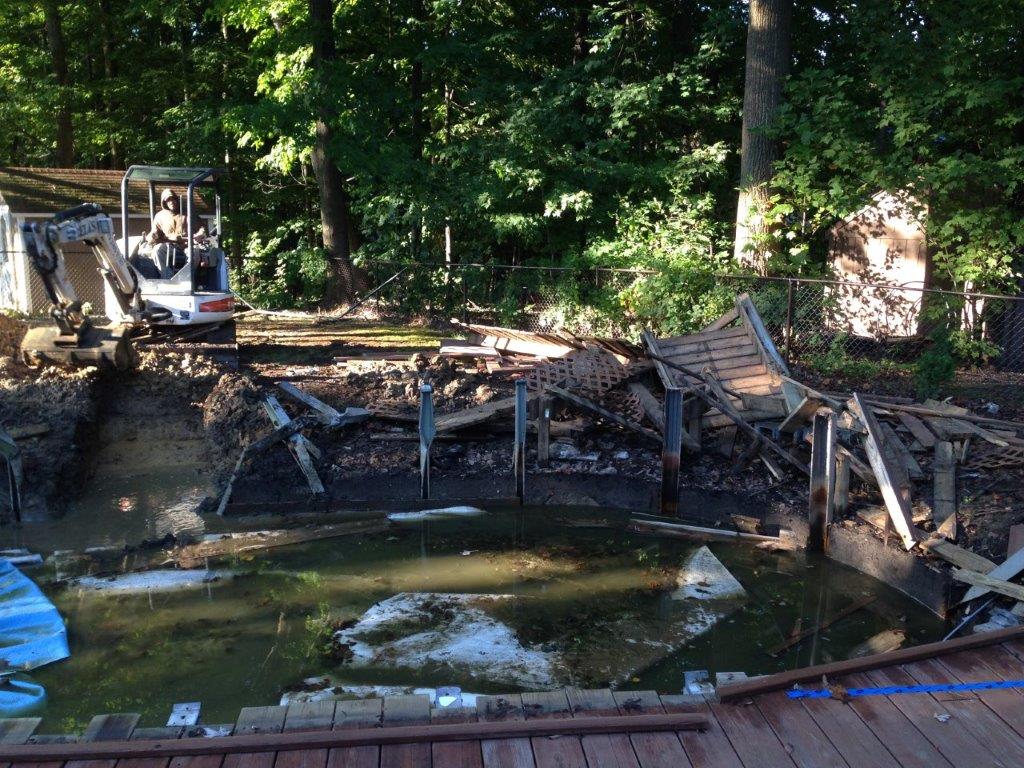Collapsed wooden bridge over a small pond in a wooded area, with debris and a small shed nearby
