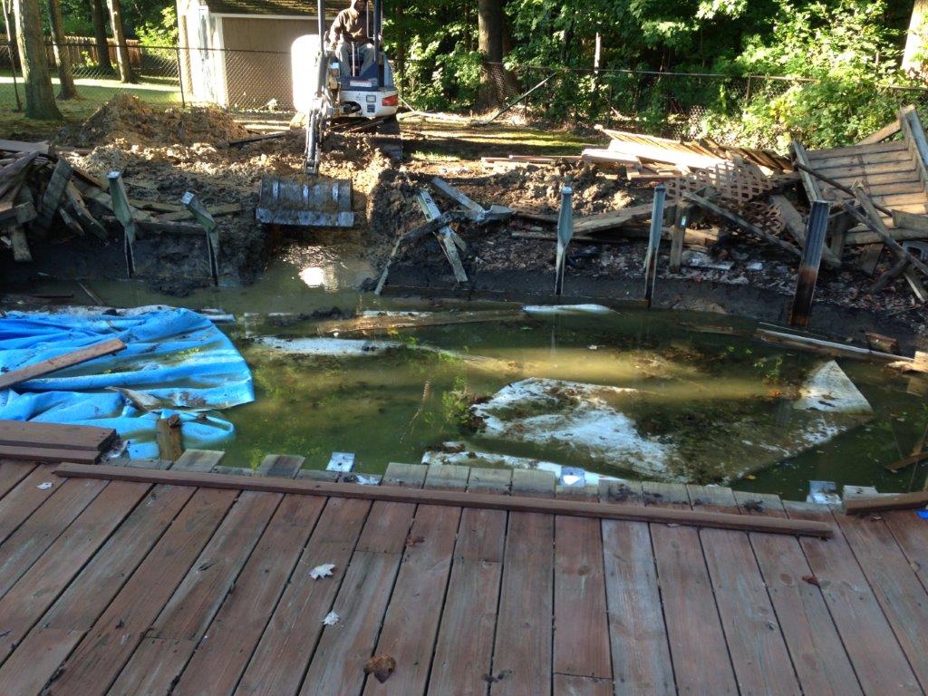 Backyard pond with murky water, blue tarp, wooden deck, and a small excavator in the background