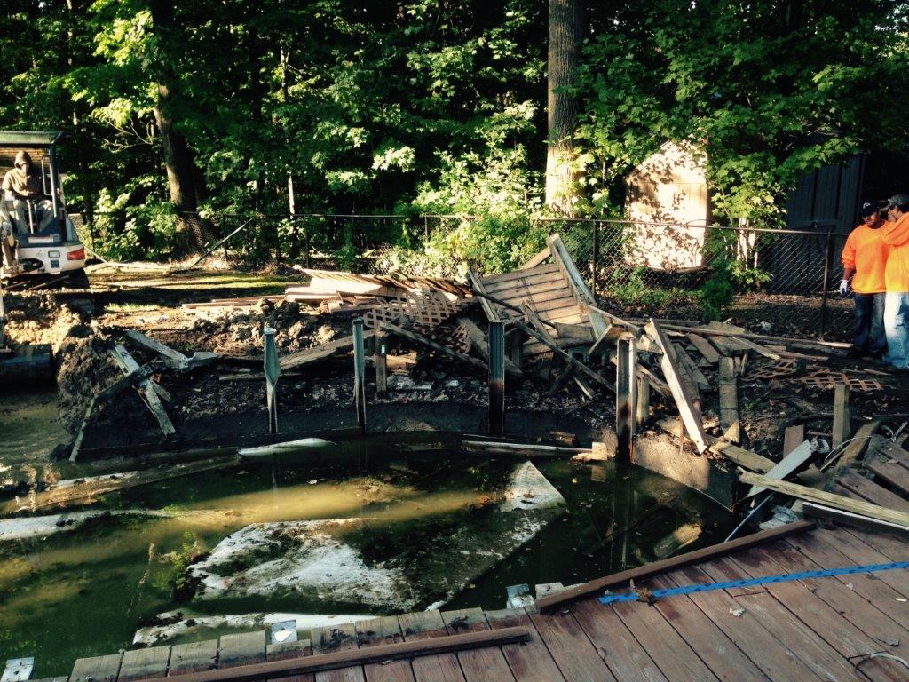 Charred remains of a burned structure beside a pond, with firefighters in orange gear in the background.