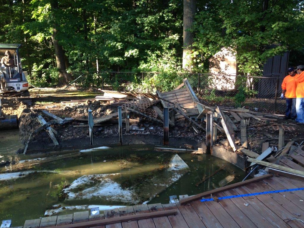 Collapsed wooden structure over a pond with debris and two workers in orange on the right