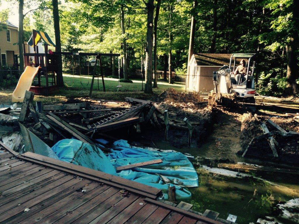 Collapsed backyard deck beside a creek, with a small excavator and a children’s playset in the trees