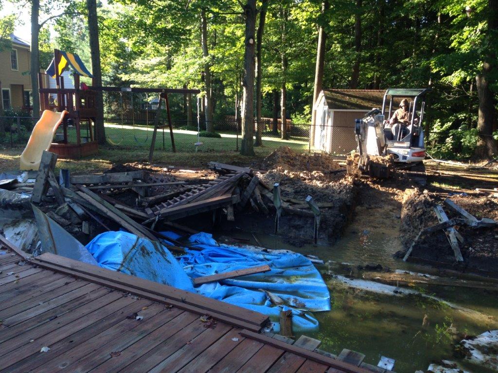 Collapsed deck by a wooded pond, with blue tarp and construction equipment nearby