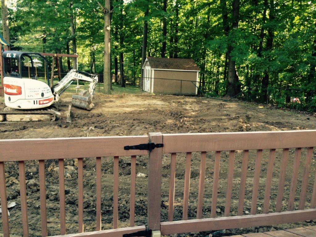 Small excavator and shed in a wooded yard behind a wooden deck railing