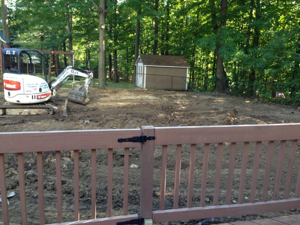 Backyard with a small excavator, dirt ground, shed, and wooded trees behind a wooden deck railing