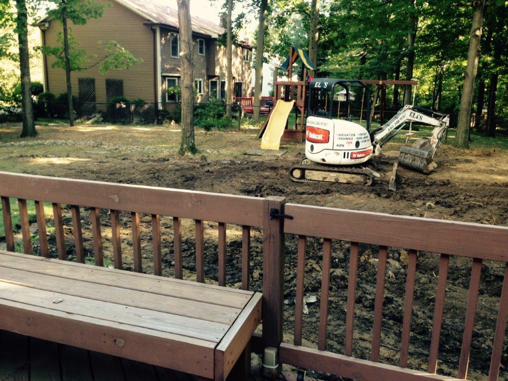 Backyard playground with a bench in front and a small excavator near a slide and playset among trees.