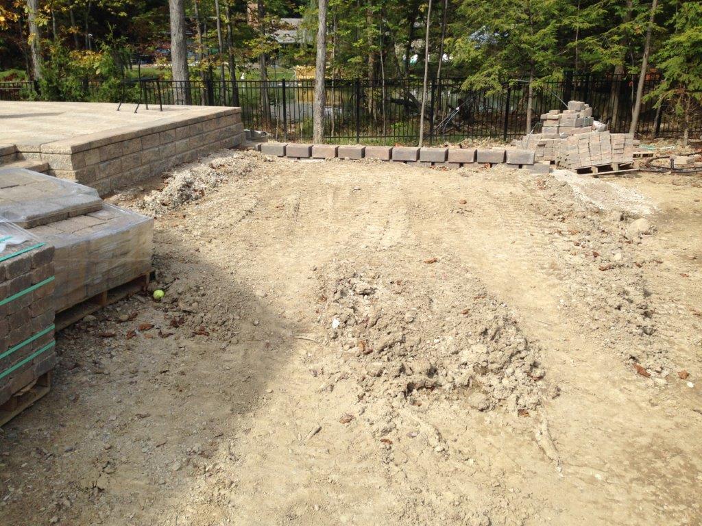 Bare dirt construction area beside stacked lumber and stone blocks, with trees in the background