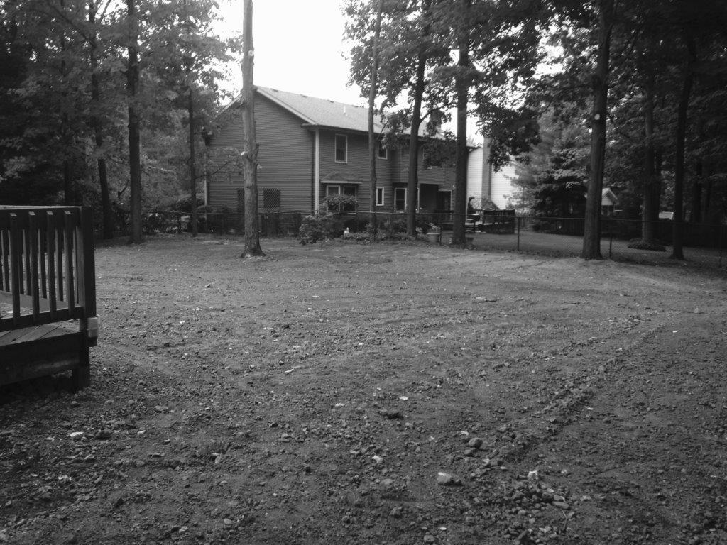 Black-and-white view of a house in a wooded yard with a deck, fence, and grassy ground