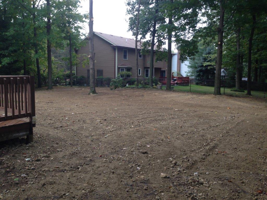 Wooded backyard with a small house, bare dirt ground, and a wooden deck on the left.