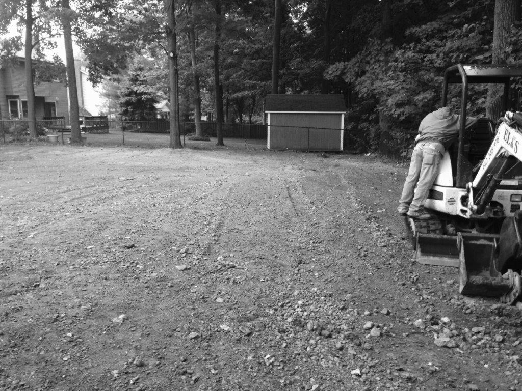 Person stands beside a backhoe in a gravel lot near trees and a small building.