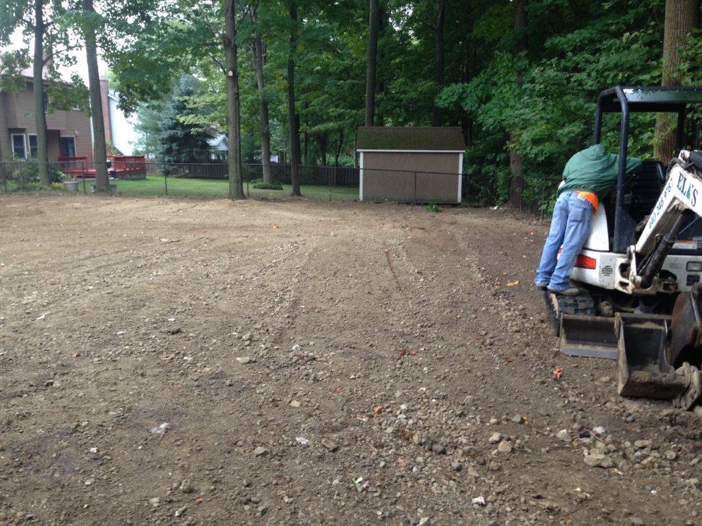Person in green jacket operating a small excavator on a dirt lot beside trees and a shed