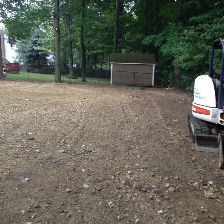 Dirt lot with trees and a small shed, with a white excavator at the right edge.