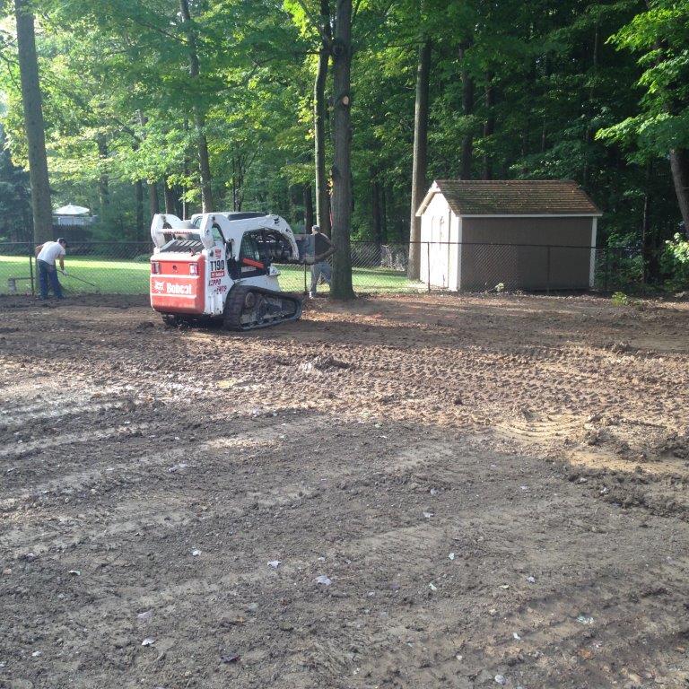 Small red-and-white skid steer on a dirt lot near a shed in a wooded park area
