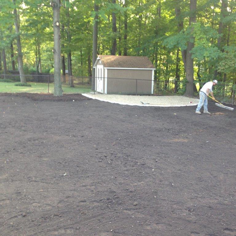 Person raking a large dirt area beside a shed in a wooded yard