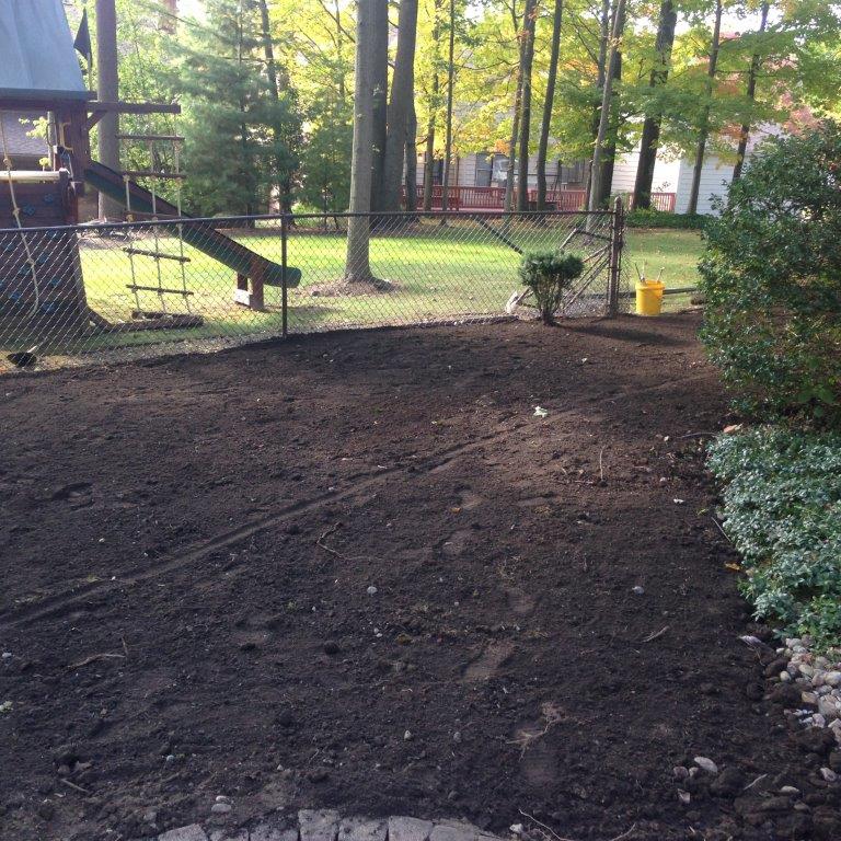 Freshly tilled soil in a fenced backyard with trees and a deck in the background